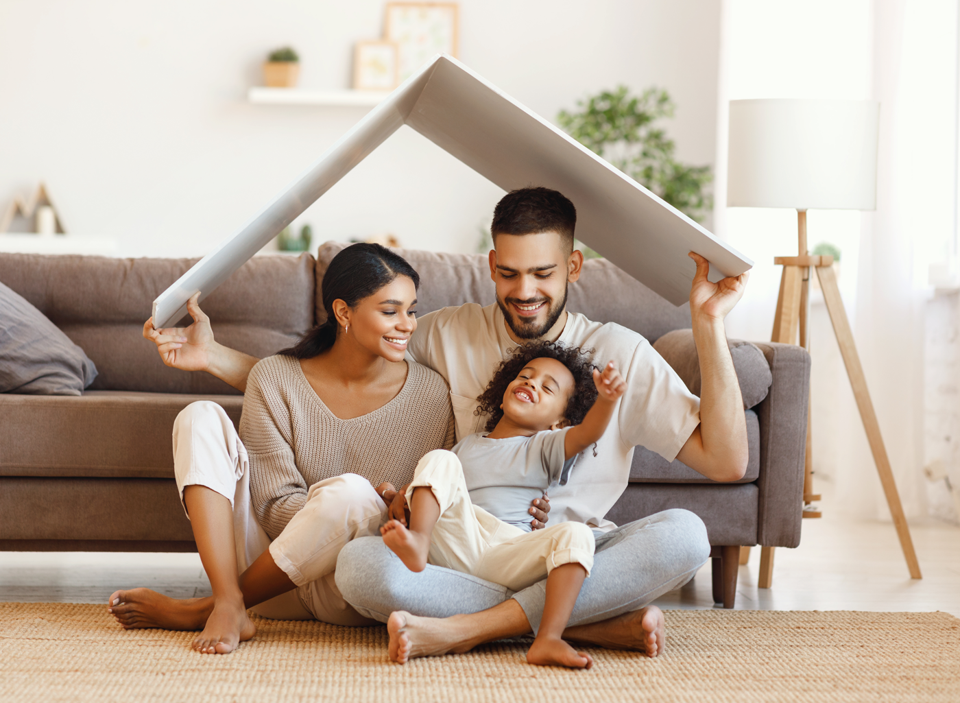 Dreaming of buying a house: a young couple sitting on the carpet in the living room of their house, the dad is carrying a cardboard roof over them